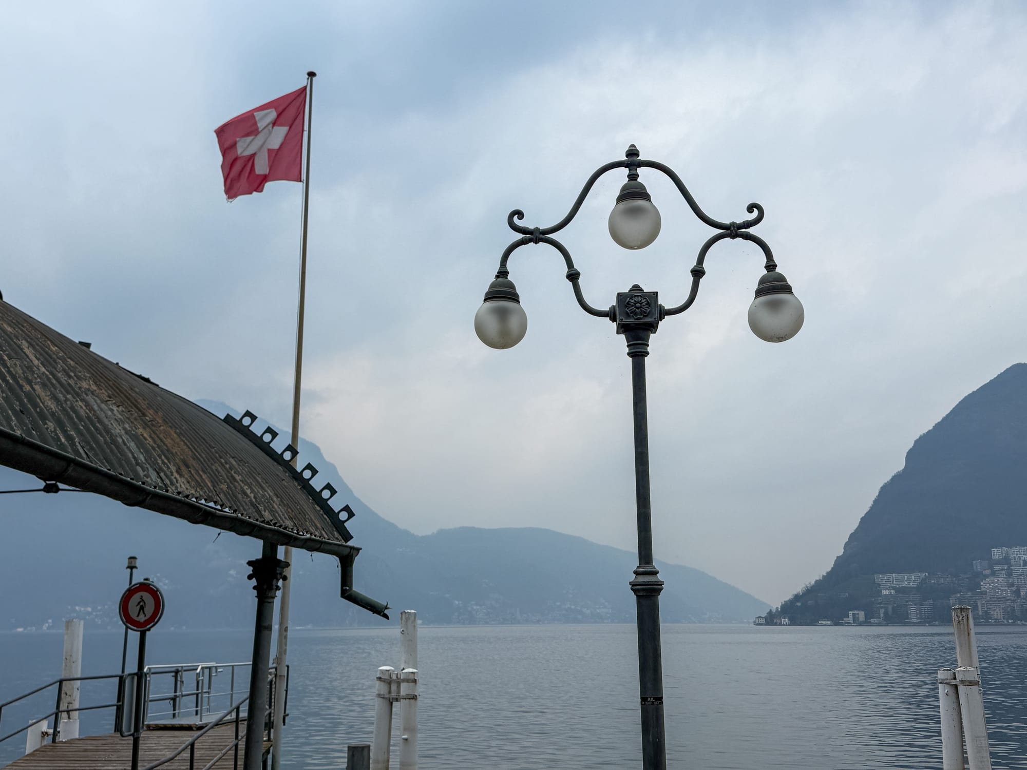 Lake Lugano (Switzerland) with a boating dock, Swiss flag, and lamp post. Campione d'Italia is visible in the background.